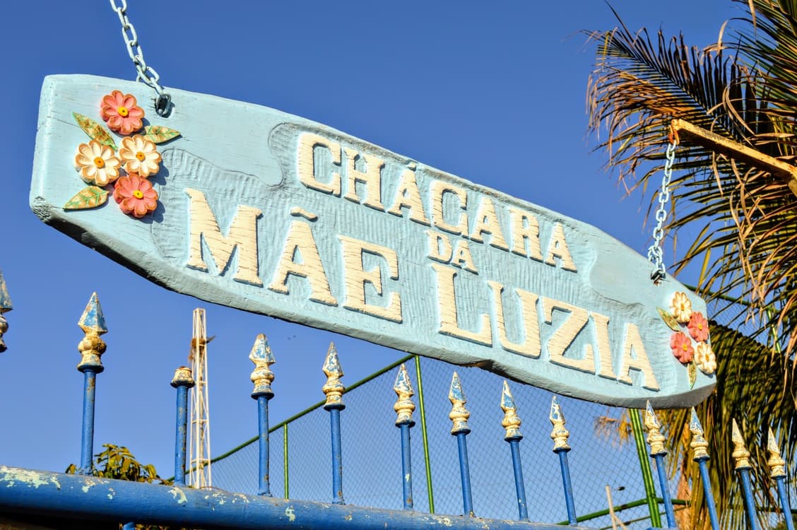 Placa de madeira pintada à mão com o nome Chácara da Mãe Luzia, cercada de flores de cerâmica.