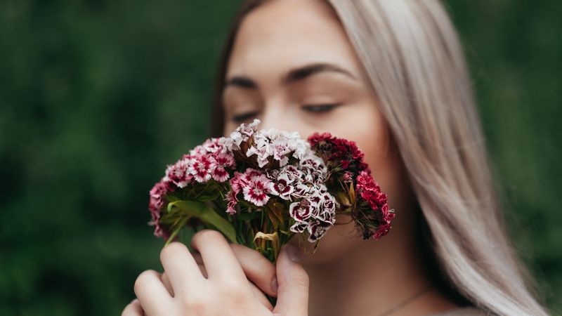 Mulher de olhos fechados aproximando o rosto de flores para sentir o aroma, cercada de folhagem verde