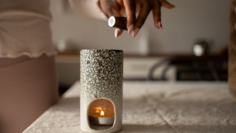 Mão pingando óleo essencial em um difusor sobre a mesa, ao lado de uma vela acesa