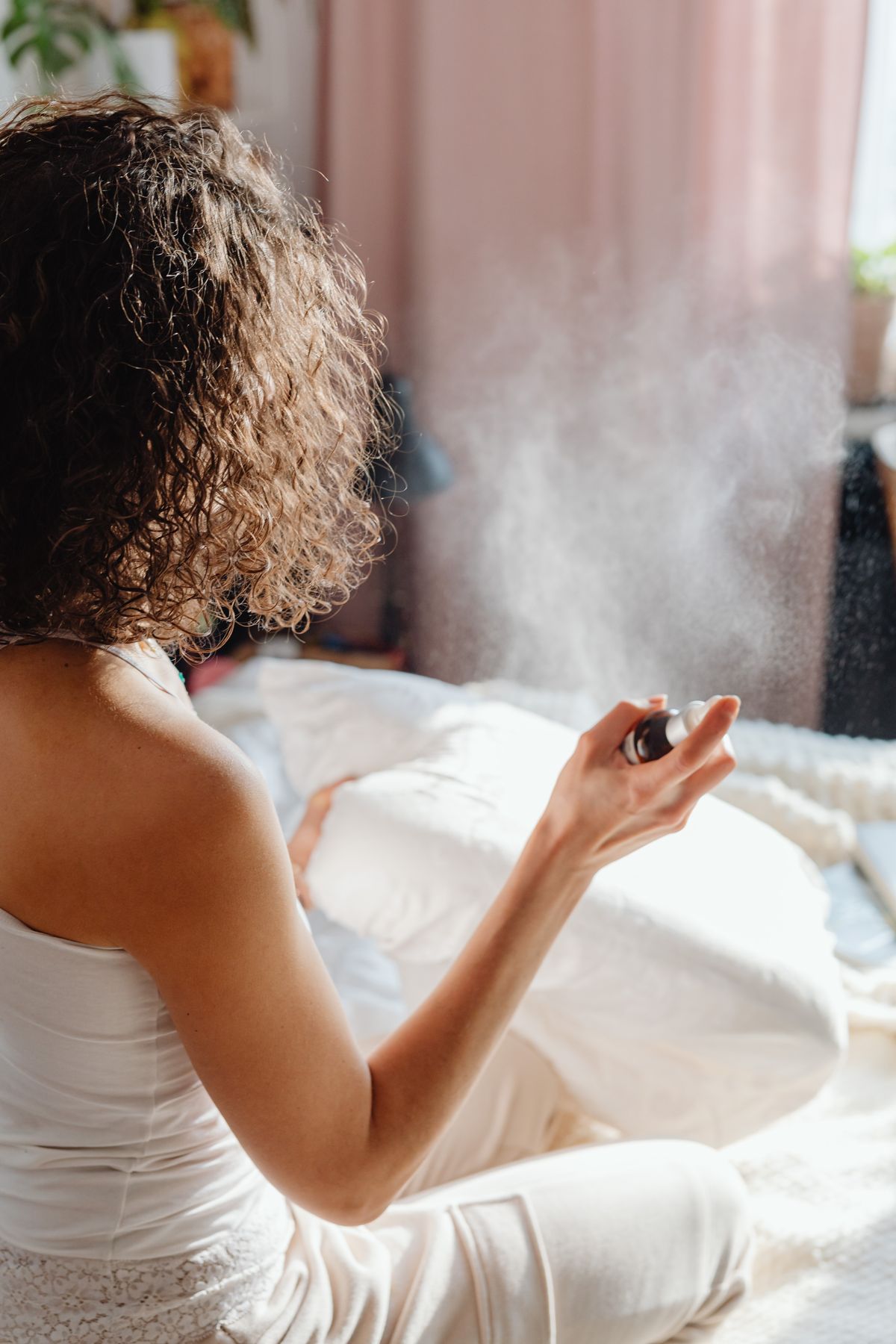 Mulher borrifando hidrolato sobre a roupa de cama num quarto com luz da manhã, a névoa fina visível contra a janela