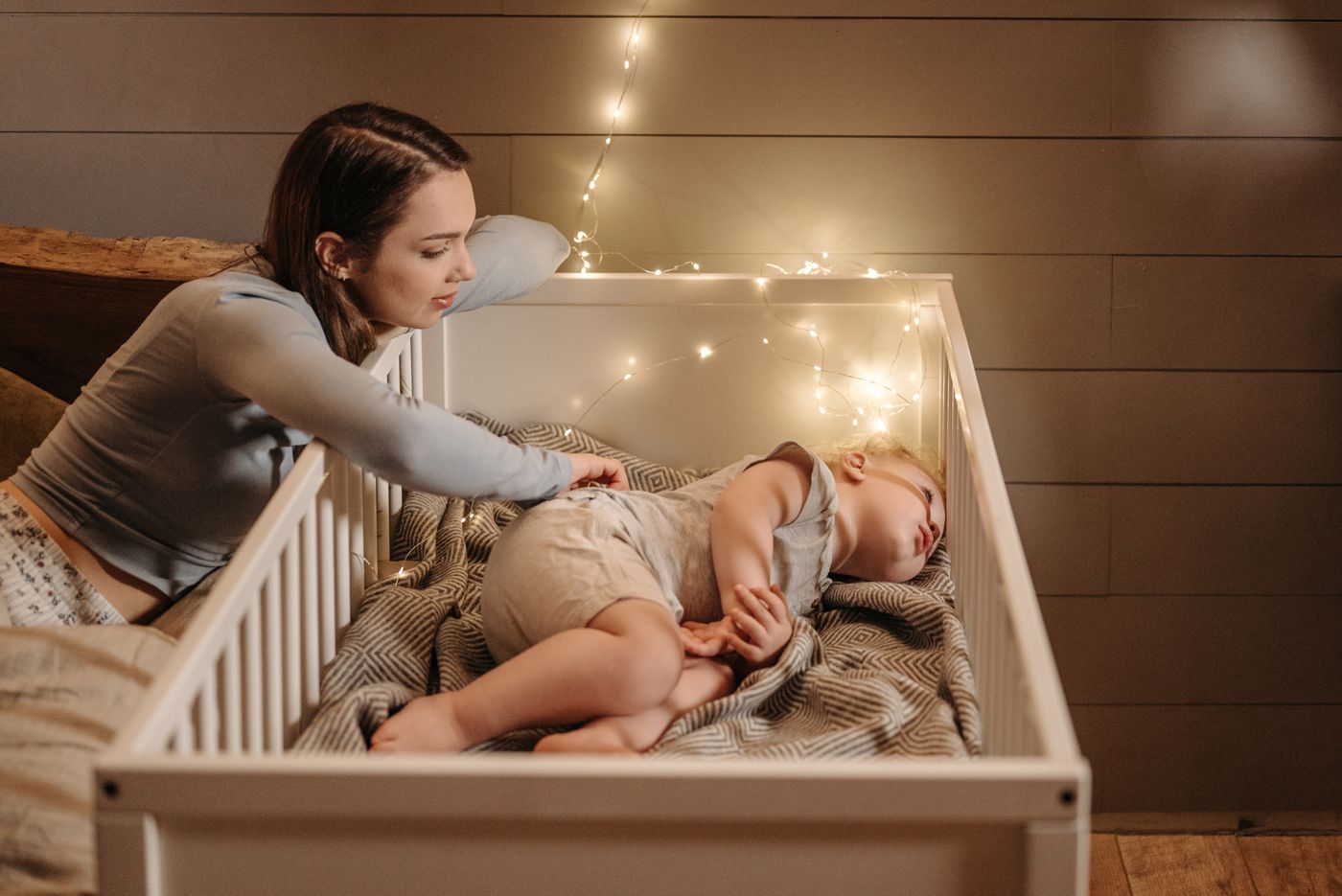 Mãe ajeitando o bebê que dorme no berço, num quarto de madeira iluminado por um cordão de luzinhas